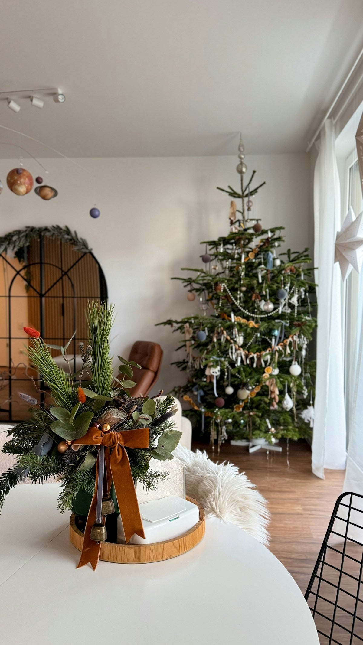 Festive evergreen arrangement with a rust-coloured velvet bow and hanging bells, with a decorated Christmas tree behind.