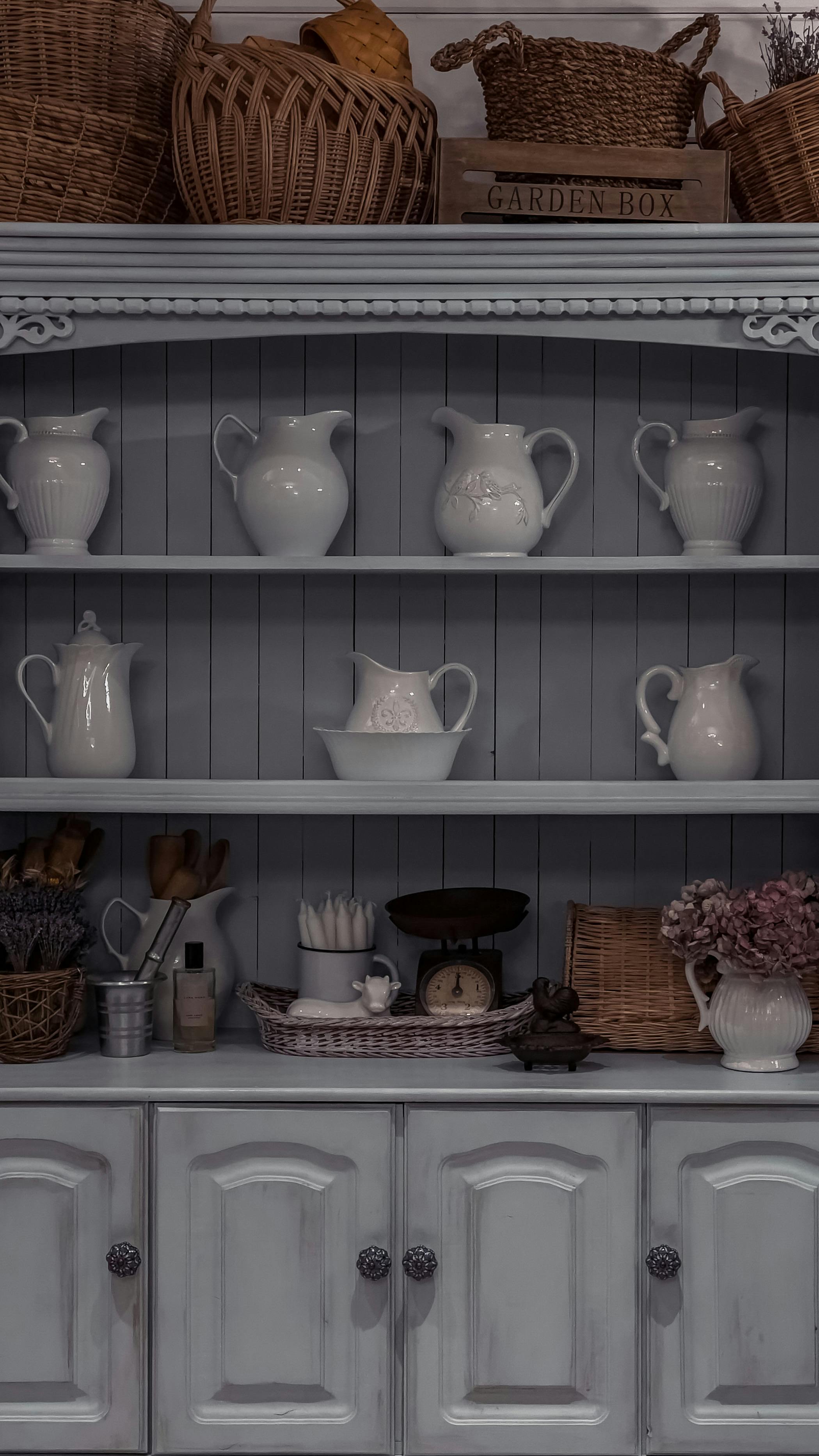 White ceramic pitchers on grey wooden shelves with wicker baskets and vintage kitchen accessories in a farmhouse-style interior.