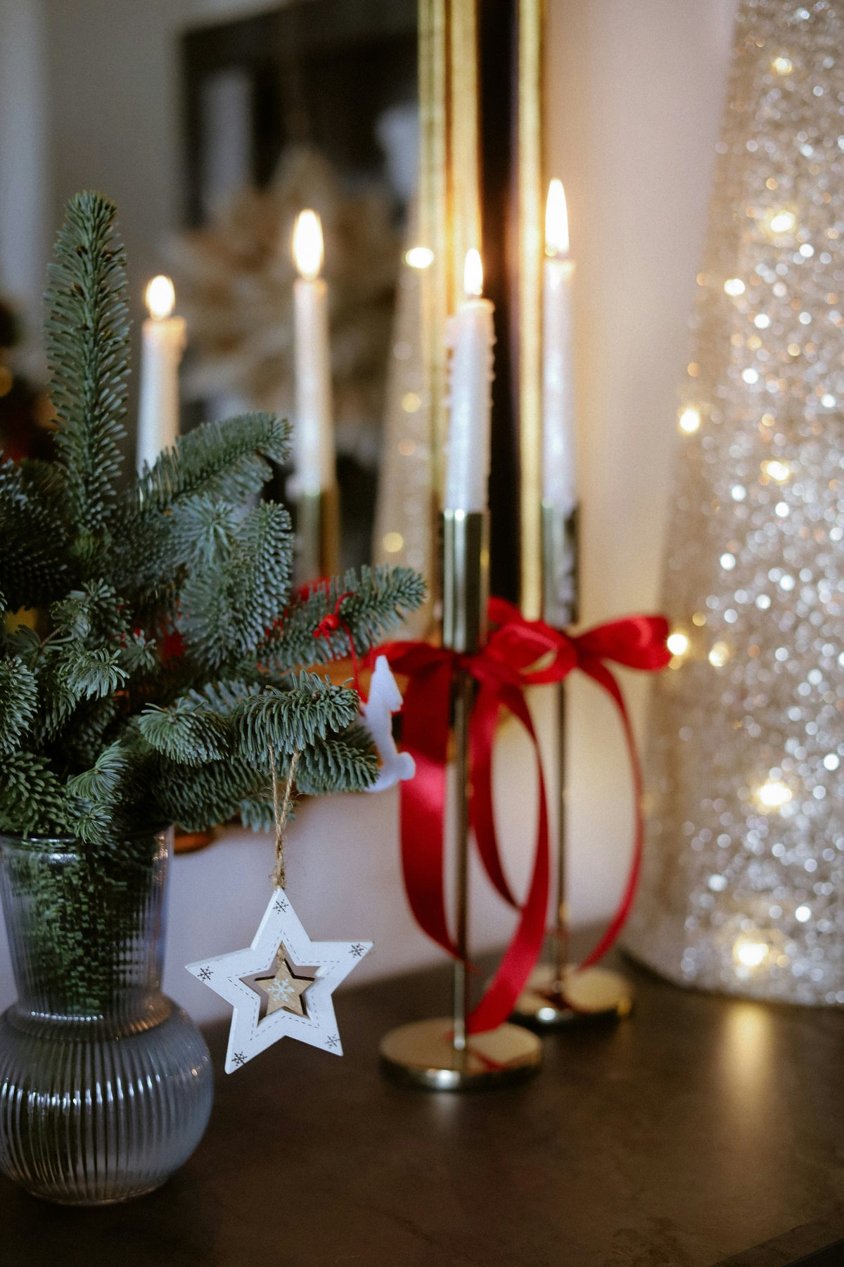 A festive holiday tabletop scene with white taper candles in gold holders, a red ribbon bow, a green pine branch arrangement in a ribbed glass vase, a white star ornament, and a glittery silver Christmas tree in the background.